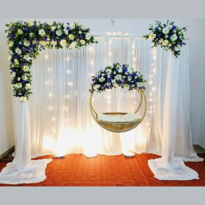Elegant white draped backdrop with fairy light drops, white chrysanthemums, blue daisy floral arrangements, suspended circular ring cradle, and red carpet for naming ceremony in Bangalore by Ease My Party