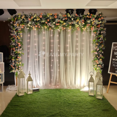 Alt Text: White galaxy netted backdrop with natural peach rose and gypsophila flower arch, fairy lights, lanterns, and green carpet matting for naming ceremony in Bangalore by Ease My Party